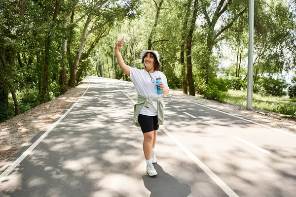 Gülümseyen genç kadın ağaç çizgili bir yolda egzersiz yapıyor, sağlıklı bir açık hava yaşam tarzını kucaklıyor..