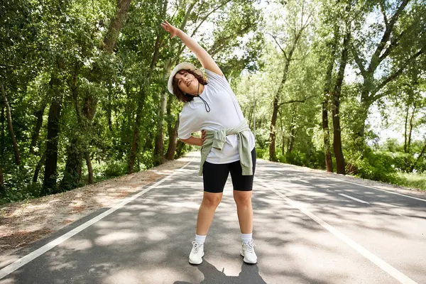 Genç ve güzel bir kadın, yemyeşil yollarla çevrili huzurlu bir yolda esneme egzersizi yapıyor..