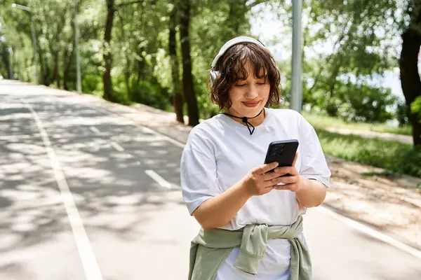 Kulaklıklı genç bir kadın yaz havasının tadını çıkarırken telefonunu doğada kontrol ediyor..
