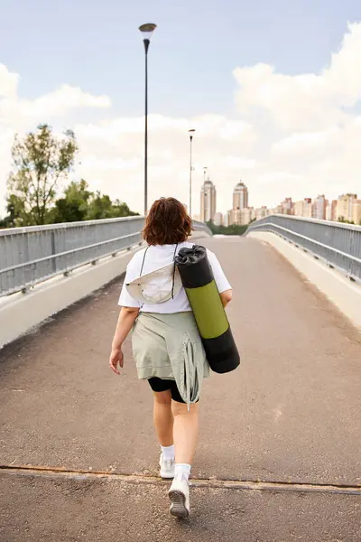 Yoga minderi taşıyan genç bir kadın güven içinde şehir köprüsünde yürür. Egzersize hazırdır..