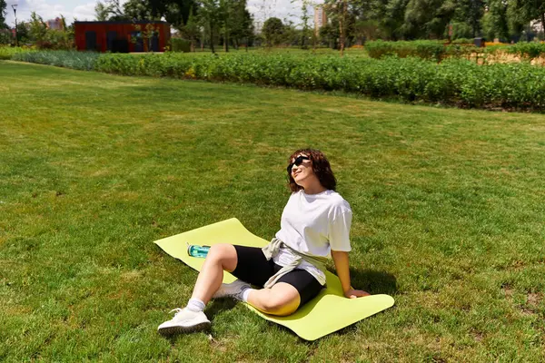 Zinde bir kadın egzersiz rutinini paspasın üzerinde yapar. Etrafı yemyeşil ve güneş ışığıyla çevrilidir..