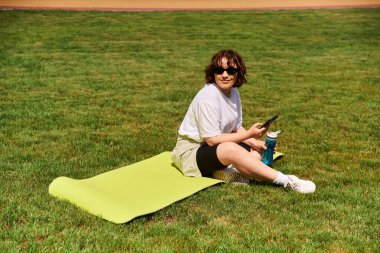 Zinde bir kadın, güneşin altındaki canlı yeşil bir parkta egzersiz yaparken minderinde mola verir..