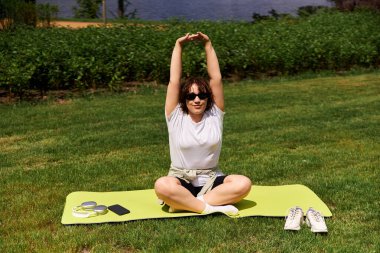 Formda genç bir kadın, canlı bir açık hava parkında yoga minderine uzanıp antrenmanının tadını çıkarıyor..