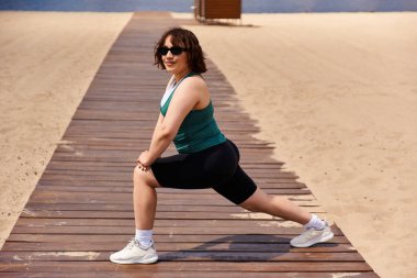 Formda genç bir kadın kumlu bir yolda hamlesini yapıyor, gün ışığında açık hava fitness seansının keyfini çıkarıyor..