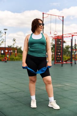 Güneşli bir günde açık hava spor salonunda direniş bandıyla esneme hareketleri yapan zinde genç bir kadın.