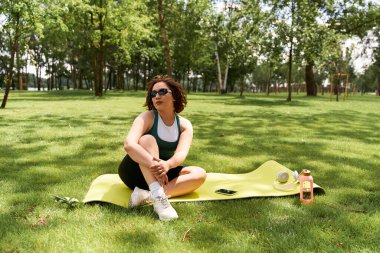 Gündelik kıyafetli genç bir kadın yemyeşil bir parktaki yoga minderinde güneşli bir günün tadını çıkarıyor..