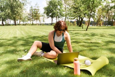 Genç bir kadın açık havada güneşli bir günün tadını çıkarıyor. Yoga minderinde dinlenirken laptopuyla çalışıyor..