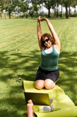 Formda genç bir kadın güneşli bir parkta yoga minderinde geriniyor ve internette spor yapıyor.