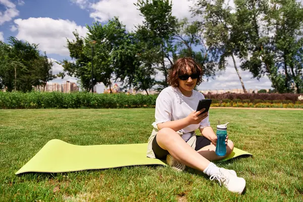Sağlıklı genç bir kadın dışarıda bir paspasın üzerinde dinleniyor, parkta spor yaptıktan sonra telefonunu kontrol ediyor..