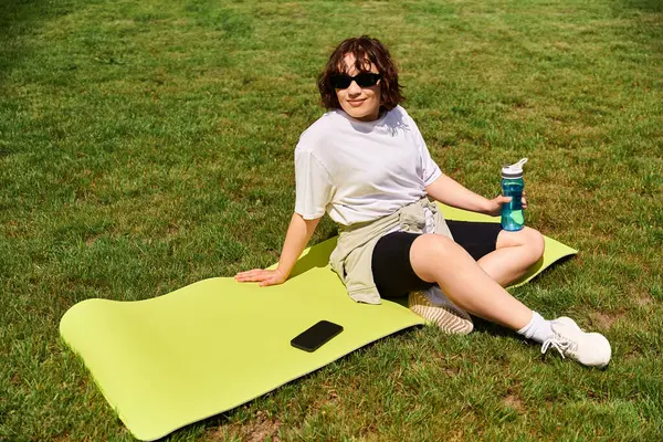 Zinde genç bir kadın yeşil bir mindere oturur, suyu yudumlar ve açık havada güneşli bir günün tadını çıkarır..