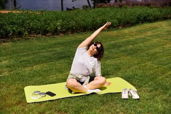 Formda bir kadın esneme hareketlerini açık havada yeşil bir paspasın üzerinde yapar..
