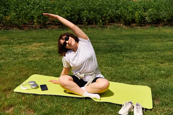 Güzel bir kadın yeşil bir paspasın üzerinde uzanır, dışarıda parkta egzersiz yaparken güneşi emer..