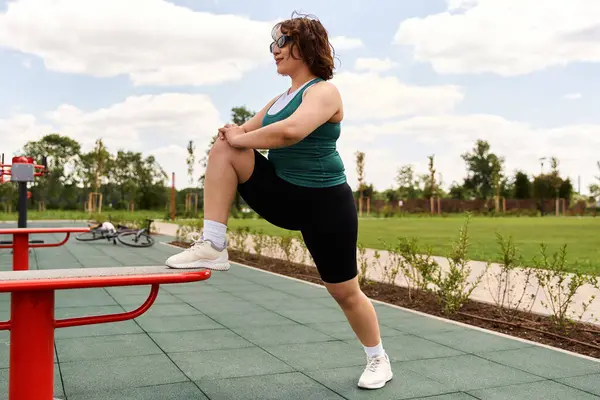 Zinde bir kadın yemyeşil bir parkta canlı kırmızı bir egzersiz istasyonunun yanında geriniyor..