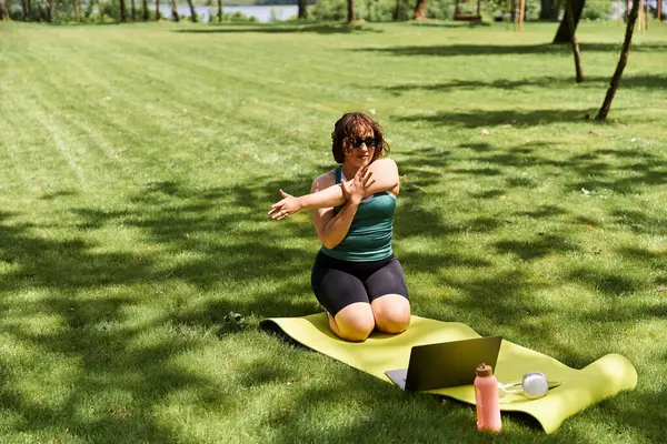 Formda ve güzel bir kadın, parkta çimenli bir alanda egzersiz yaparken esniyor ve internetteki fitness 'i destekliyor.