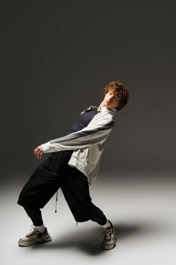 Kıvırcık saçlı genç bir adam minimalist stüdyo ortamında cesur dans hareketleri sergiliyor..