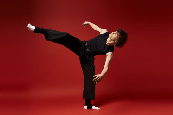 Curly young man in black attire performs an energetic dance against a bold red backdrop.