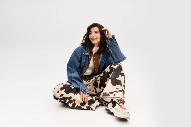 Tarz sahibi genç bir kadın, gri bir arkaplan karşısında, modaya uygun bir inek deseninin içinde gelişigüzel oturuyor..