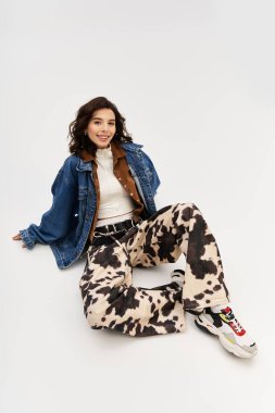 İnek desenli pantolon ve kot ceket giyen genç bir kadın, gri bir zemine karşı neşeli bir şekilde poz veriyor..