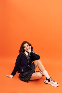 Genç bayan düşünceli bir şekilde oturuyor, bir stüdyo ortamında şık bir ceket ve etek giyiyor..