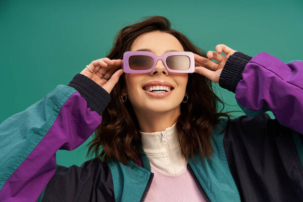 A confident young woman in trendy sportswear poses with sunglasses against a turquoise backdrop.