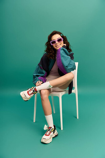 A fashionable young woman in sporty attire poses confidently on a chair in a studio setting.