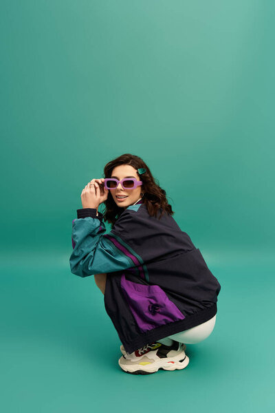 A young woman in a trendy sporty outfit poses playfully in a studio with a turquoise background.