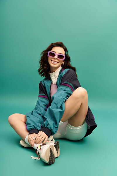 A young woman showcases her fashionable sporty look while sitting on a turquoise background.