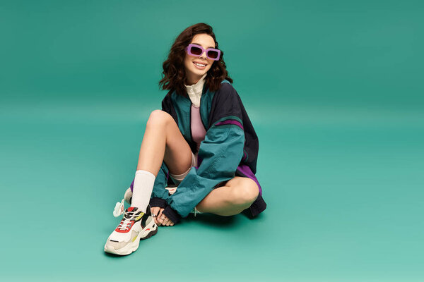 Sporty young woman in a trendy outfit relaxes confidently against a vibrant turquoise backdrop.