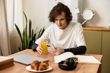 Rahat giyimli genç bir adam evde rahatlıyor, meyve suyunu yudumluyor ve akıllı telefonunu kullanıyor..