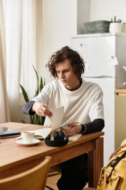 Genç adam, rahat bir evde kahvesini yudumlarken bir kitaba dalmış, ahşap bir masada oturuyor..