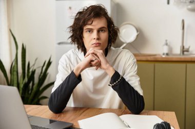 Genç bir adam, rahat giyinmiş, rahat bir evde, ahşap bir masada düşünceli bir şekilde oturur..