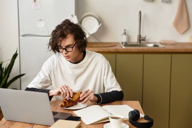 Genç bir adam mutfak masasında oturur, derslerine ve bilgisayarına odaklanırken kruvasanın tadını çıkarır..