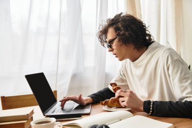 Genç bir adam rahat bir evde kruvasanın tadını çıkarırken dizüstü bilgisayarıyla çalışıyor..