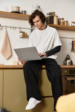 Rahat giysiler içinde genç bir adam mutfak tezgahına oturur ve dizüstü bilgisayarına odaklanır..
