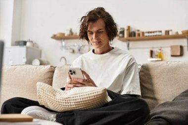 Genç bir adam rahat bir şekilde kanepede oturuyor, akıllı telefonuna dalmış, rahat bir ev kıyafeti giyiyor..