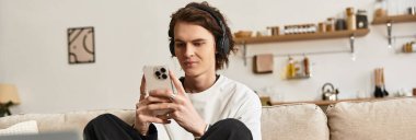 Genç bir adam rahat giysiler içinde evinde rahatlıyor, akıllı telefonuna odaklanıyor..
