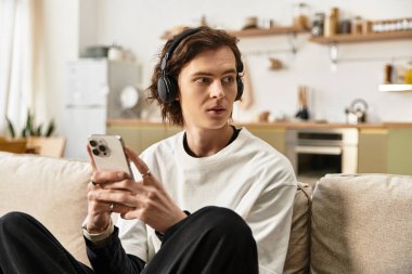 Genç bir adam, şık kulaklıklarıyla akıllı telefonuna odaklanmış bir şekilde kanepede dinleniyor..