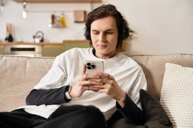 Genç adam rahat bir şekilde kanepede oturuyor, akıllı telefonuna dalmış, rahat ve rahat giysiler giyiyor..
