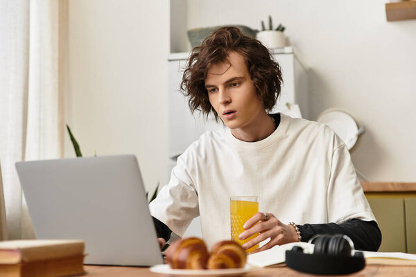 Молодой человек в удобной одежде играет со своим ноутбуком, потягивая сок за завтраком.