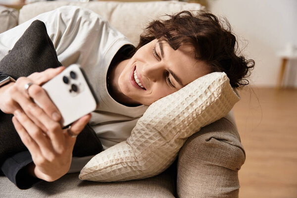 Young man lies comfortably on a sofa, smiling while engaged with his smartphone, feeling cozy.
