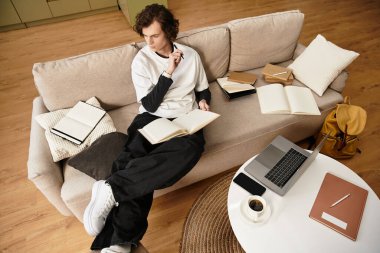 Samimi giyimli genç bir adam, bir kanepeye oturur. Çalışma odasına dalmış, etrafında defterler ve bir dizüstü bilgisayarla..