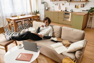 Genç adam rahat bir şekilde kanepede uzanıyor, evde kitap okumaya ve dinlenmeye odaklanıyor..