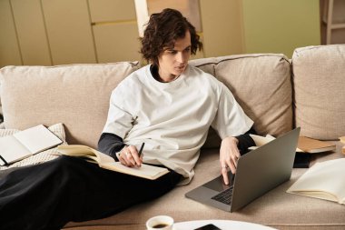 Genç bir adam evdeki bir kanepede rahatça oturur, elinde dizüstü bilgisayar ve defterle ders çalışır..