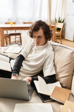 Rahat giyinen odaklanmış genç adam evde açık kitapların arasında dizüstü bilgisayarla meşgul oluyor..