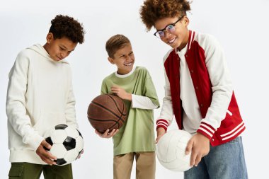 Three adorable boys share joy while holding basketball, football, and volleyball in trendy wear.