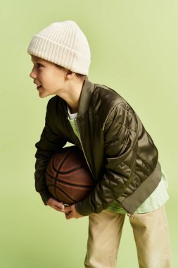 Moda kıyafetleri içinde yakışıklı bir çocuk basketbol topu tutuyor ve eğlenceli bir ortamda gülümsüyor..