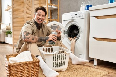 Gülümseyen genç adam evde rahatlıyor, çamaşır sepetini katlıyor ve çamaşır makinesini yakınlarda tutuyor..