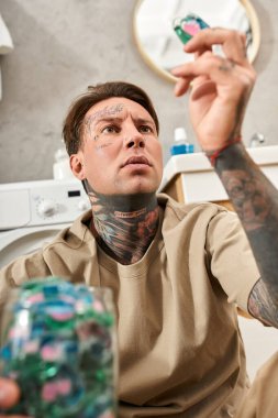 Genç, dövmeli bir adam rahat bir şekilde evinde oturur, rahat bir ortamda çamaşır kapsüllerine odaklanır..