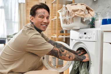 Genç dövmeli adam evde rahat bir anın tadını çıkarırken çamaşır makinesini mutlu bir şekilde dolduruyor..