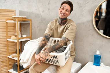 Dövmeleri olan genç bir adam evinde çamaşır sepetini tutarken sevinçle gülümsüyor..